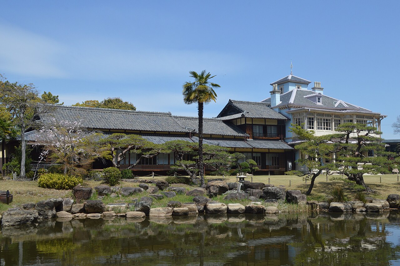 A pale-blue Western-style mansion with a turret and grey tiled roof rises behind a long Japanese-style tiled house, seen across a garden pond with rock edging, trimmed pines, a palm tree and a stone lantern under a blue sky.