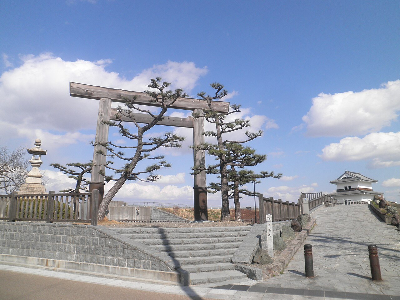 A tall plain wooden torii gate stands among twisted pines and a stone lantern at the top of stone steps, with a small white-walled tower and a stone-paved path to the right under a blue sky with clouds.