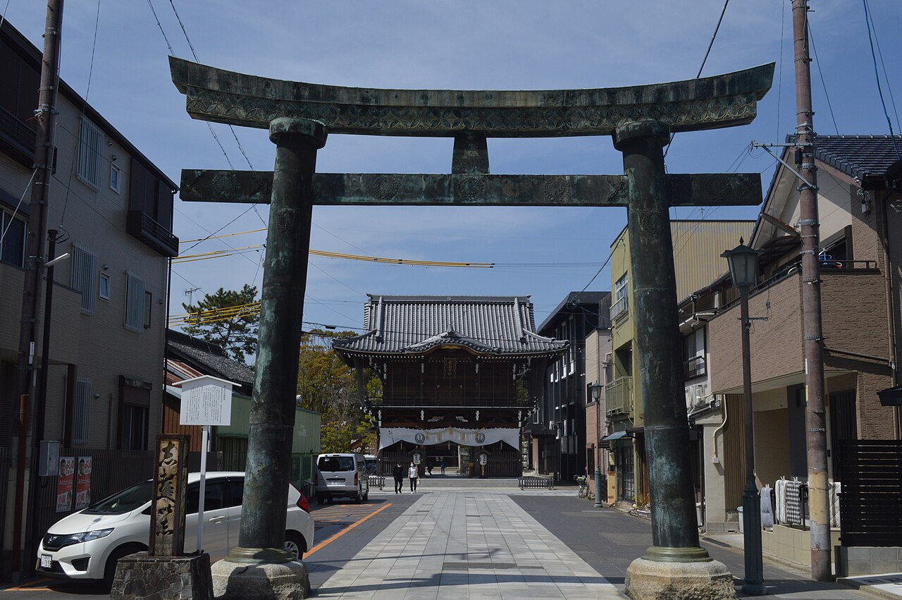 A weathered bronze torii gate with green patina stands over a narrow street of houses and a parked car, leading the eye to the tiled two-storey gate of a shrine hung with white cloth curtains.