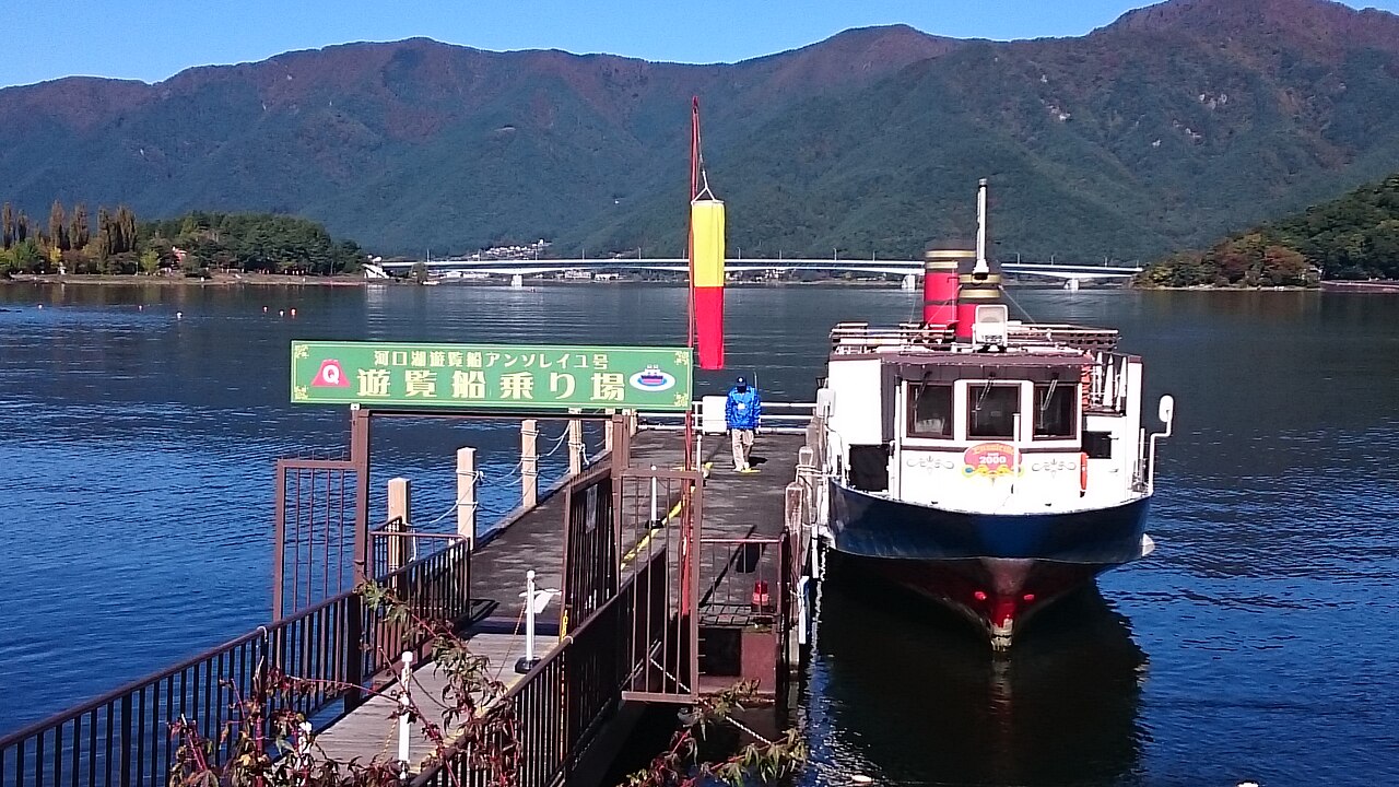 A lake sightseeing boat moored at a wooden pier on Lake Kawaguchi, with wooded hillsides rising on the far shore.