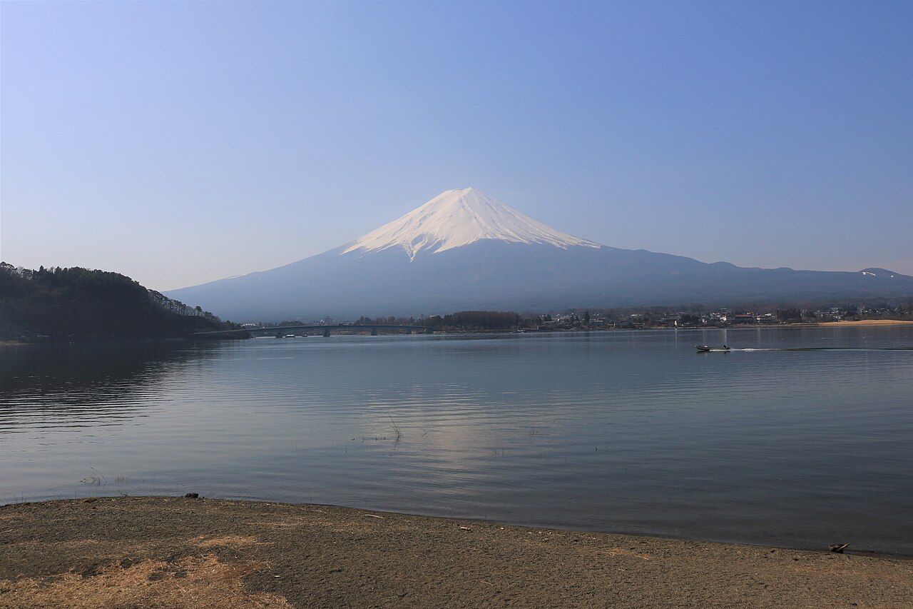 Snow-capped Mount Fuji rising beyond the calm water of Lake Kawaguchi, seen from a sandy stretch of the shore.