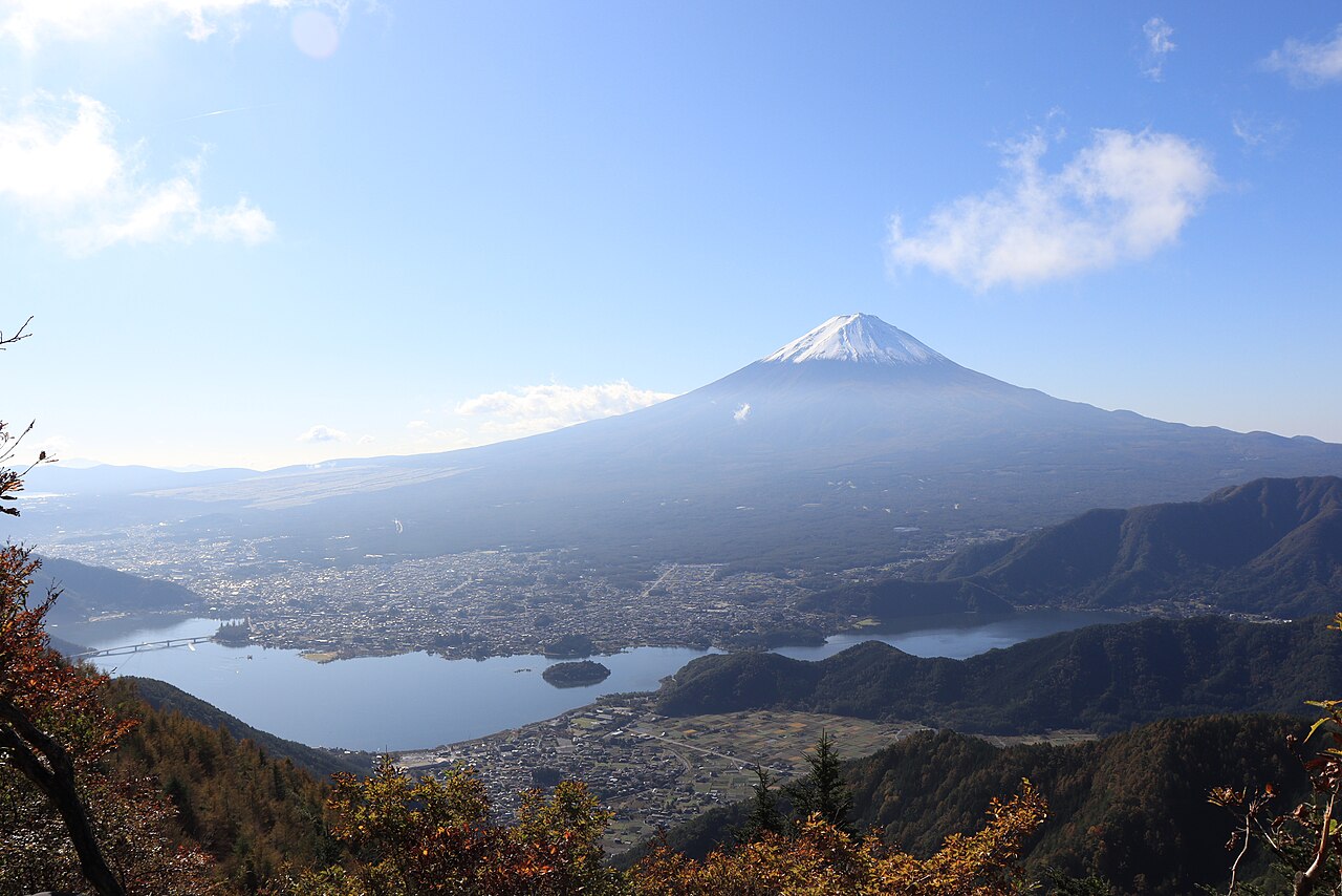 An elevated view over Lake Kawaguchi and the lakeside town, with Mount Fuji and layered mountain ridges filling the background.