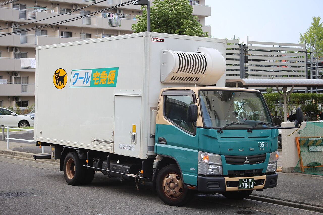 A white refrigerated Yamato Transport delivery truck with a teal cab and the black cat logo beside the Cool TA-Q-BIN sign, parked on a street