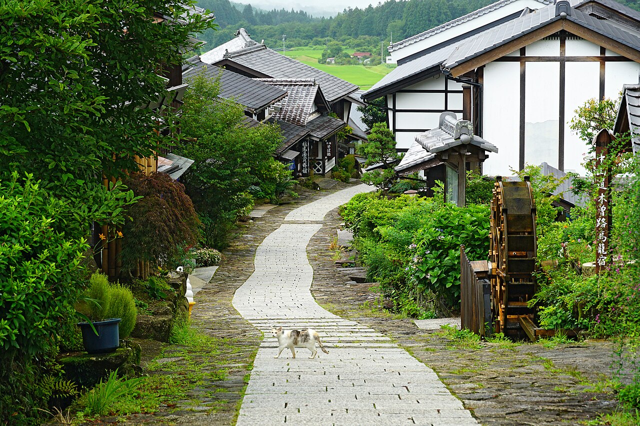 A pale stone-paved lane curving uphill through Magome-juku between green gardens and old wooden houses, with a small waterwheel beside the path