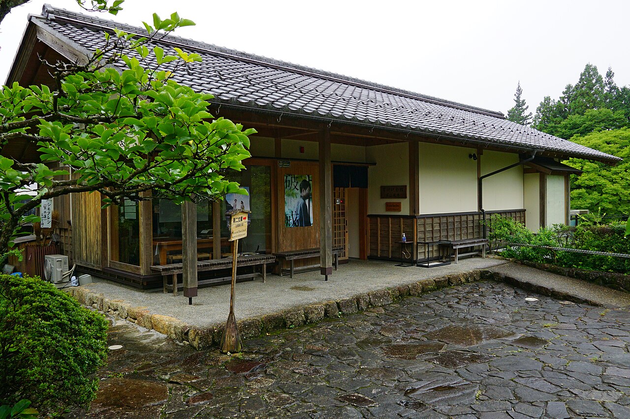 A low white-walled museum building with a tiled roof and a wooden veranda, set on a cobbled forecourt among trees