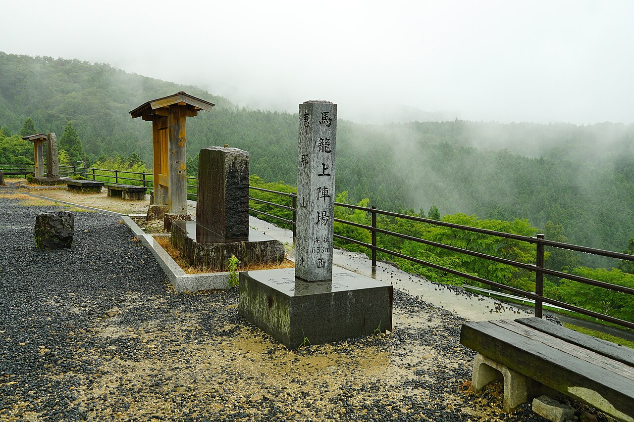 Stone monuments and a small wooden-roofed marker at a railed overlook above a valley half-hidden in low cloud, with a bench at the side