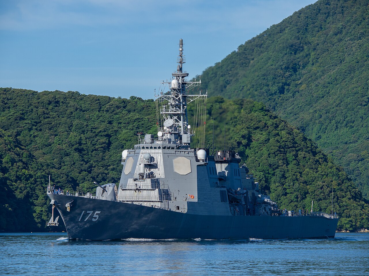 A grey Aegis destroyer moves through blue water in front of forested hills on Maizuru Bay