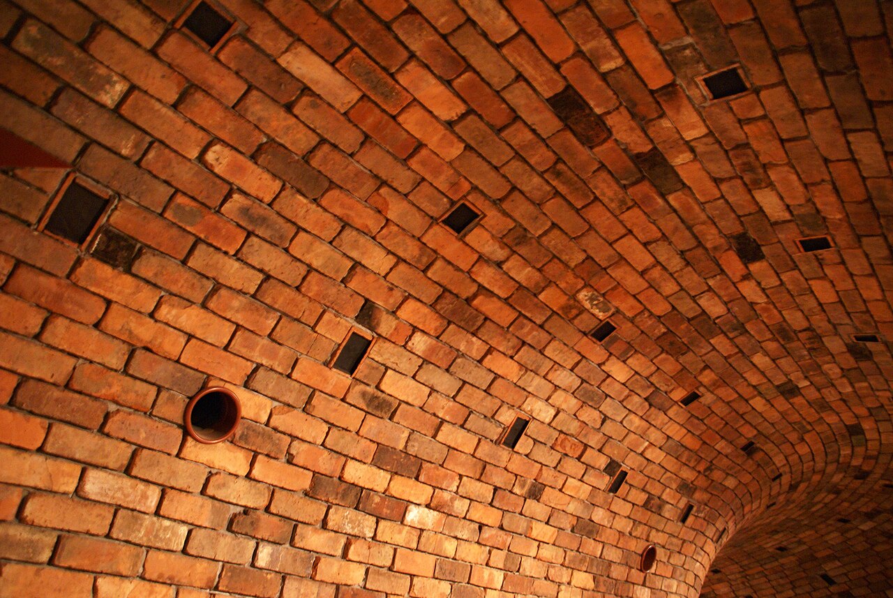The curved brick-lined ceiling of a vaulted interior, with small dark vent openings and a round pipe end set into orange brickwork