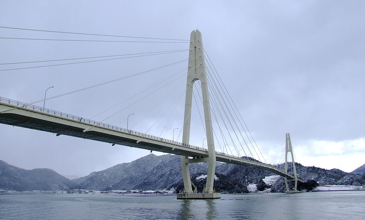 A white cable-stayed bridge on tall slender pylons crosses a grey bay under a heavy winter sky, with hills dusted in white behind