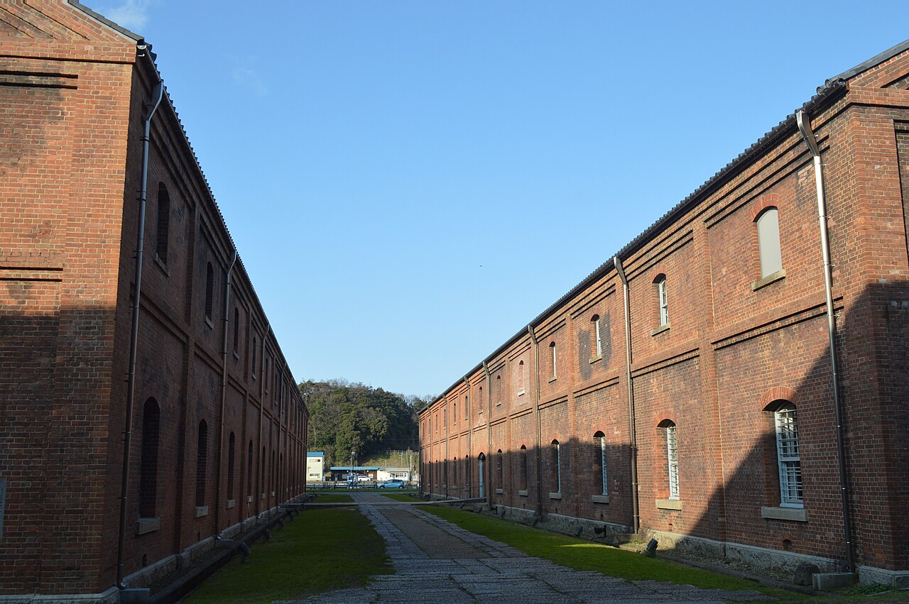 Two long two-storey red brick warehouses face each other across a narrow stone-paved lane with grass edges, under a clear blue sky, with wooded hills at the far end