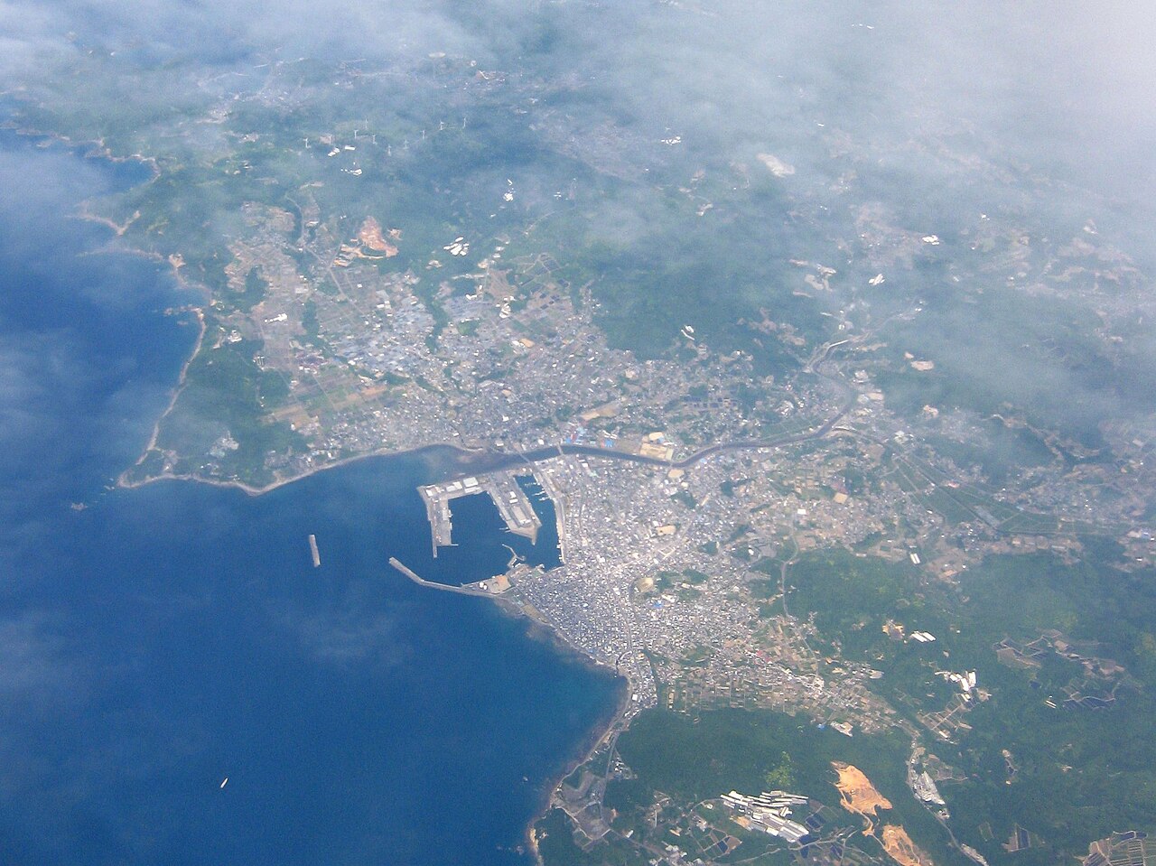 An aerial view of a coastal town packed around a rectangular harbor with long breakwaters, with deep blue sea on the left and green hills under thin cloud.
