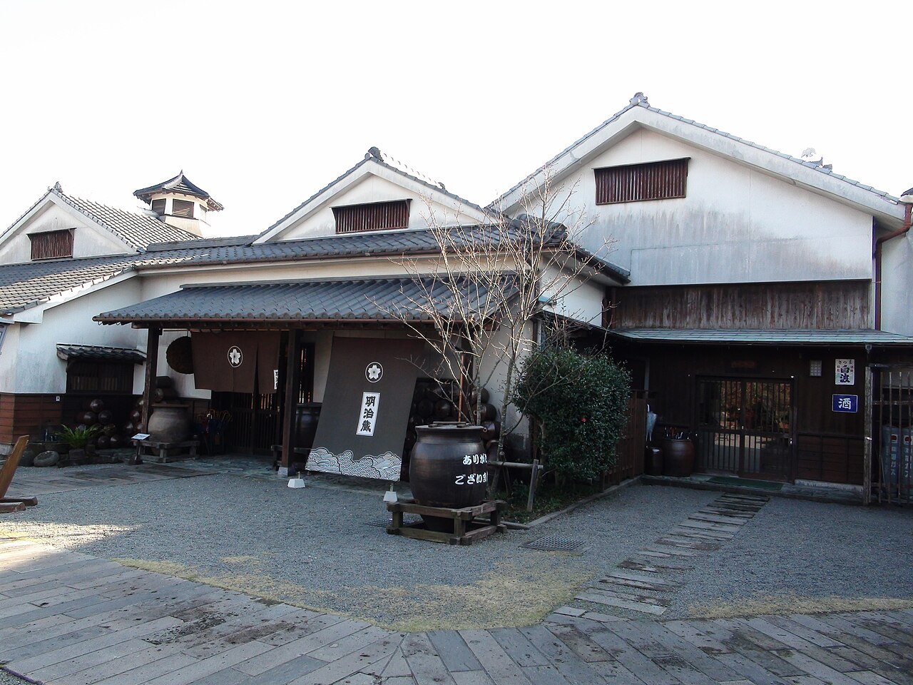 Low white-walled storehouses with dark tiled roofs and a wooden gate, a large brown pot and a dark noren curtain on a gravel courtyard.