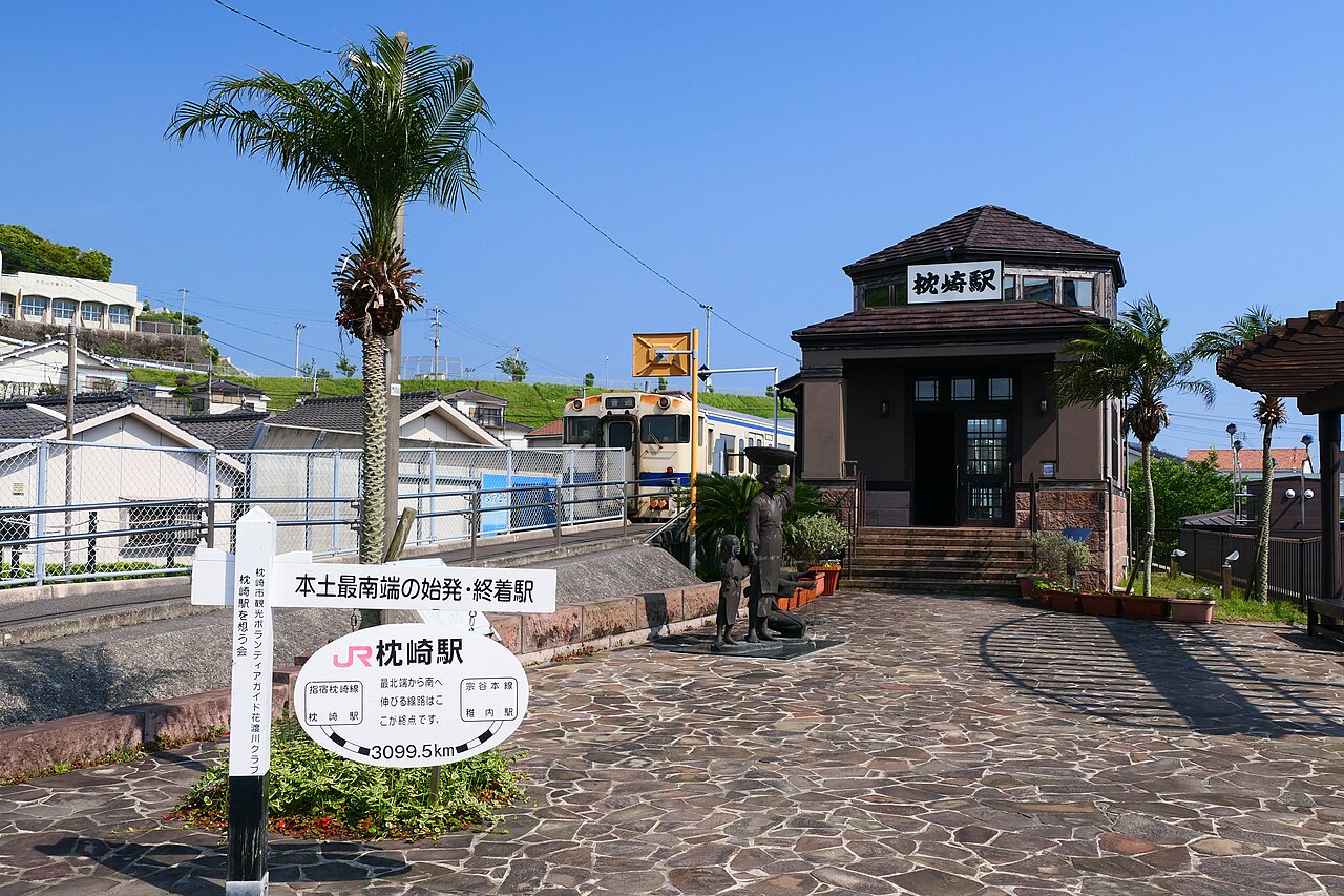 A small brown station building with a clock and a station-name sign stands above a stone-paved square with a bronze statue of a woman and child, a yellow-and-white train waiting behind a fence, under a clear blue sky.