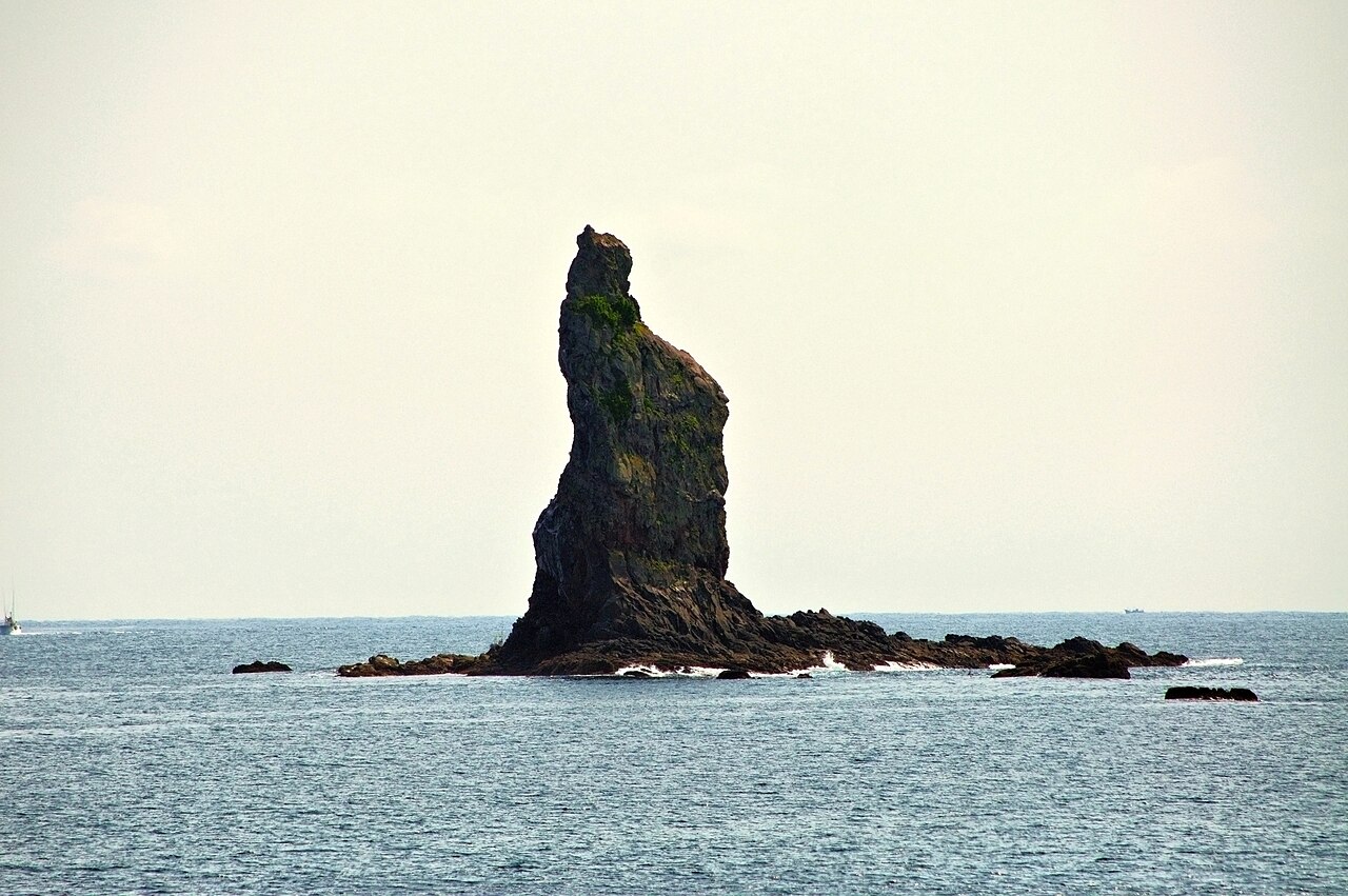 A tall, narrow dark rock stack rising straight out of a calm blue sea with small waves breaking at its base and a pale hazy sky behind.