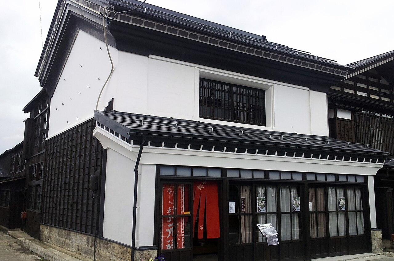 A two-story storefront with white plaster walls, a black timber lower half and a tiled roof, with a red noren curtain at the entrance.
