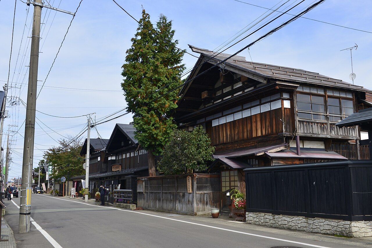 A quiet street lined with old dark-timber merchant houses, black plank fences and a stone base wall, with utility poles and trees under a blue sky.