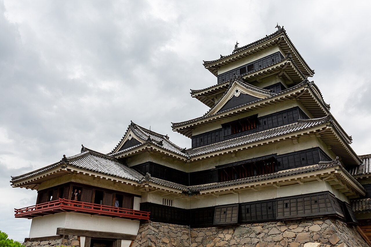 Close view of Matsumoto Castle's multi-tiered keep with its black wooden walls and layered gabled roofs, seen from below with a red-railed turret at left.