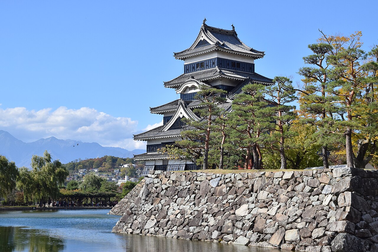 Matsumoto Castle's black-and-white keep rising behind a pine-topped stone wall, with the moat in the foreground and mountains beyond under a clear blue sky.
