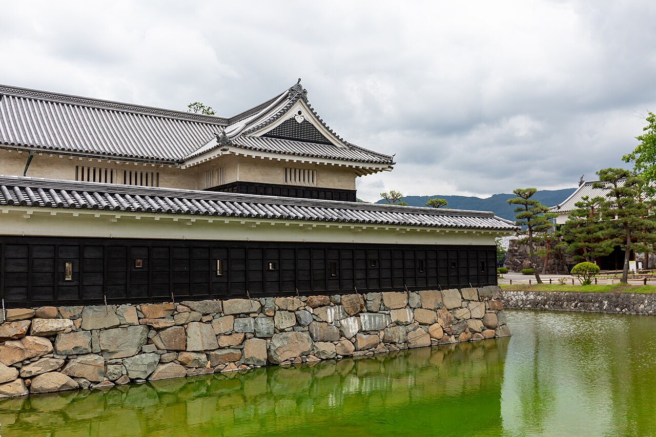 A black-boarded castle turret wall on a stone base at the edge of a green moat in Matsumoto, with pine trees and mountains in the distance.