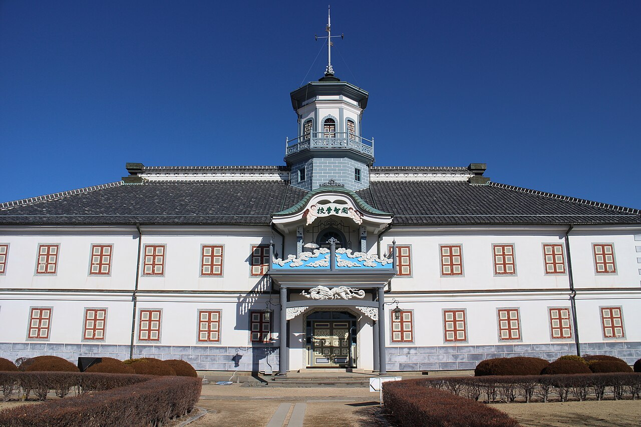 Front of the Former Kaichi School in Matsumoto, a white Meiji-era school building with reddish window frames, an octagonal cupola and ornate blue-and-white carving over the entrance under a deep blue sky.