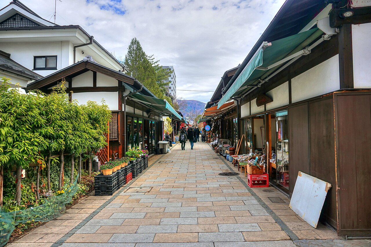 Narrow stone-paved Nawate-dori lane in Matsumoto lined with small wooden shops and green awnings, with shoppers walking toward distant mountains.