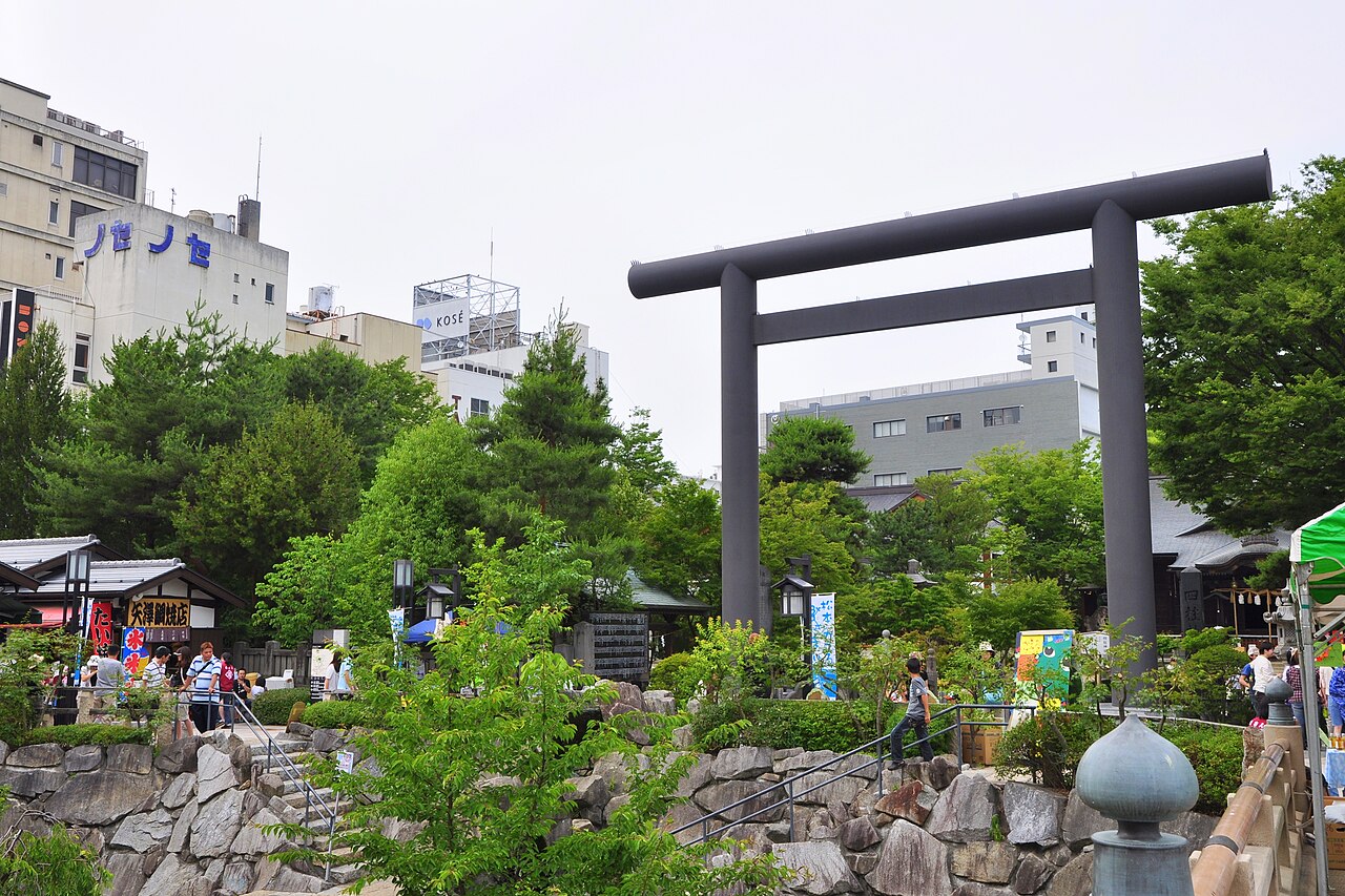 Large dark torii gate at Yohashira Shrine in central Matsumoto, rising above stone-walled planting beds with visitors and city buildings behind the trees.