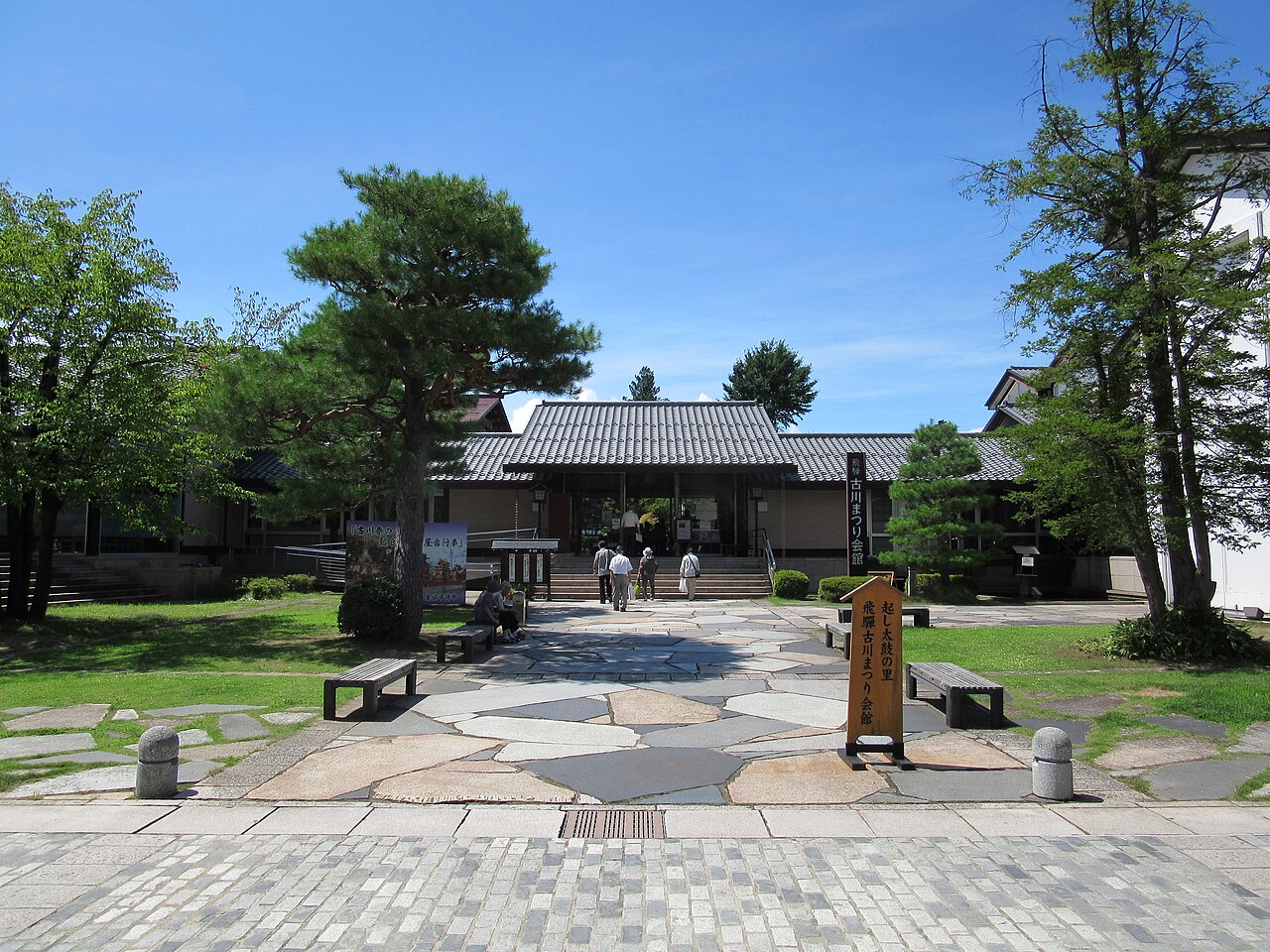 The forecourt and entrance of Hida Furukawa Matsuri Kaikan, with stone paving, pine trees, a wooden sign and a few visitors in the distance under a blue sky