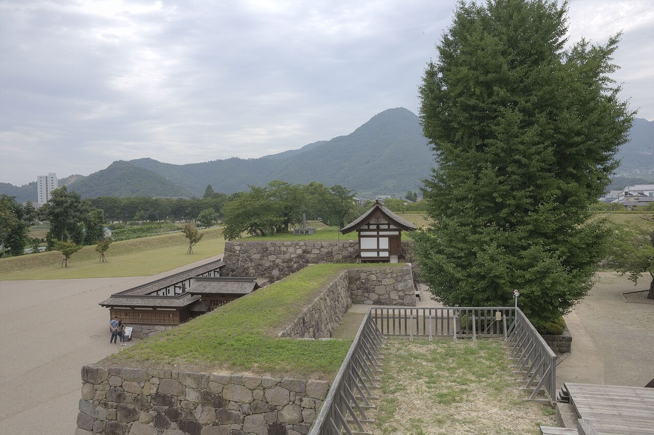 The open inner bailey of Matsushiro Castle ruins with low stone walls, a tall conifer and a small gate tower, framed by green mountains.