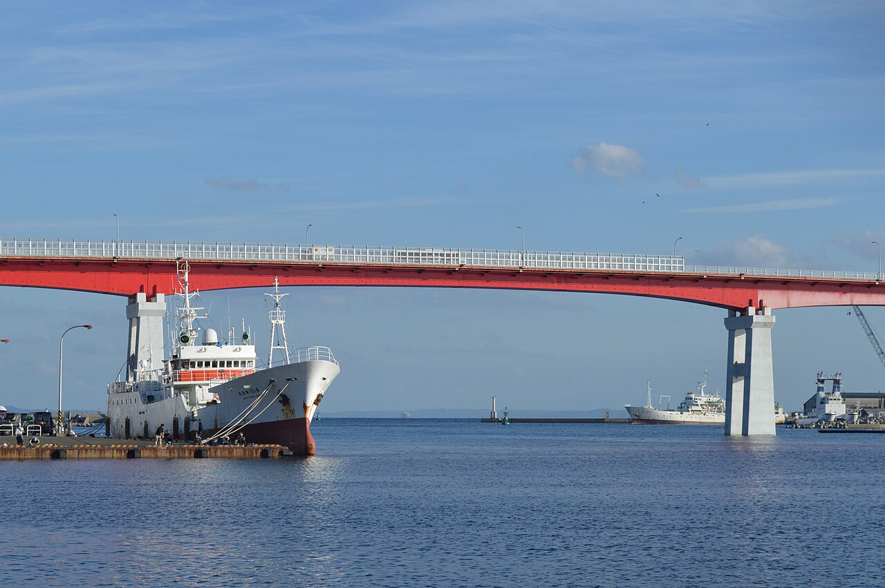 A moored ship in Misaki harbor in front of the long red Jogashima Bridge crossing the blue water under a bright sky