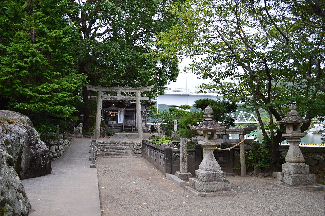A small shrine behind a stone torii gate under large trees, with stone lanterns in the foreground and a bridge over the river in the distance