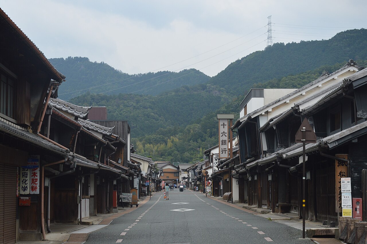 Street view of Mino's Udatsu townscape preservation district, with two-story machiya wooden houses lining a paved street toward wooded hills.