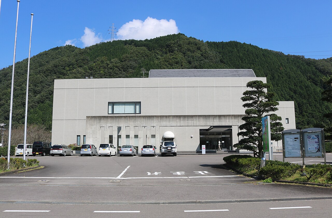 Exterior of the Mino Washi no Sato Kaikan museum building, with a parking area and wooded hillside behind it.