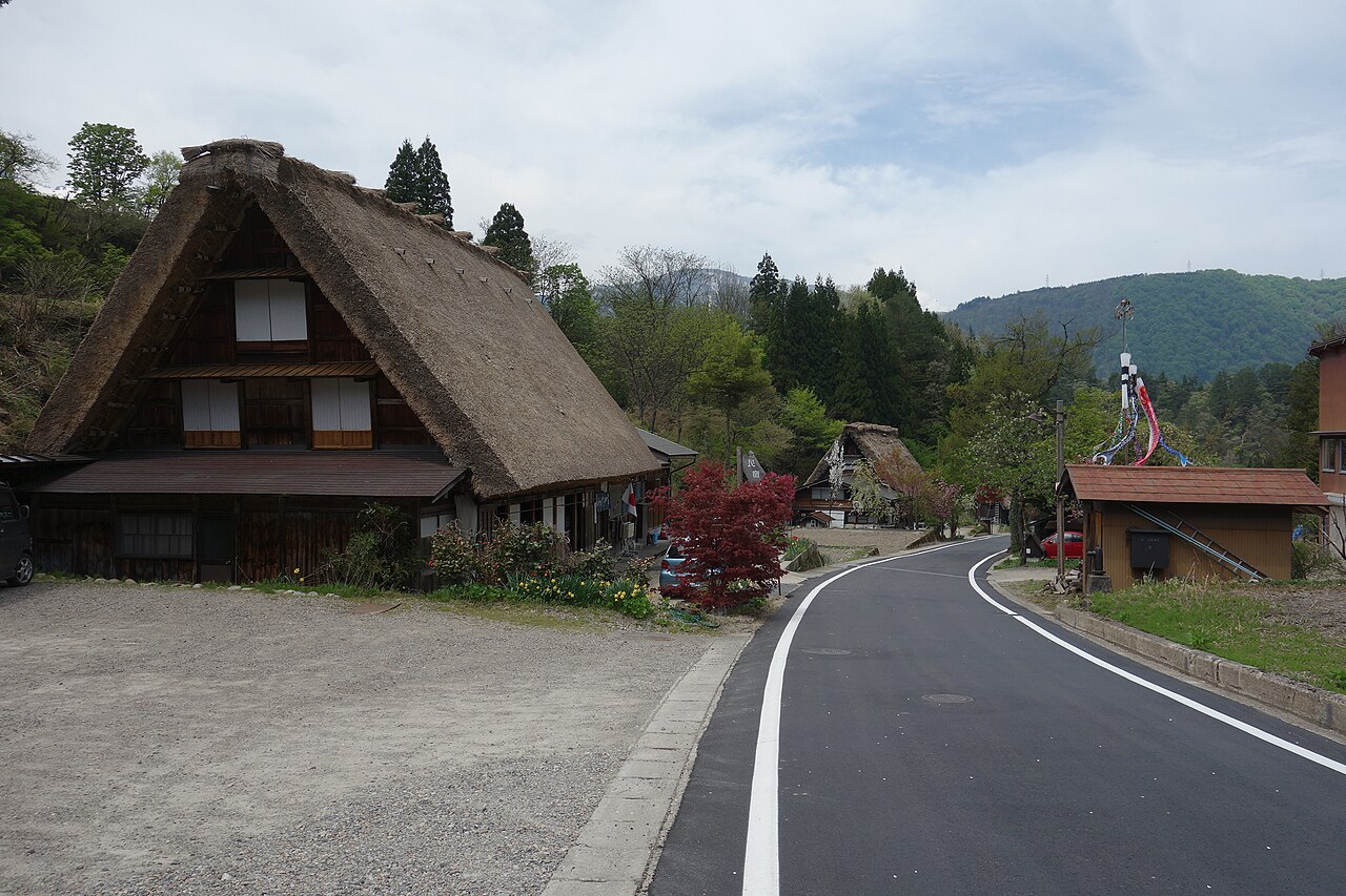 A thatched gassho-style farmhouse listed as a minshuku beside a road in Shirakawa village, with forested hills behind