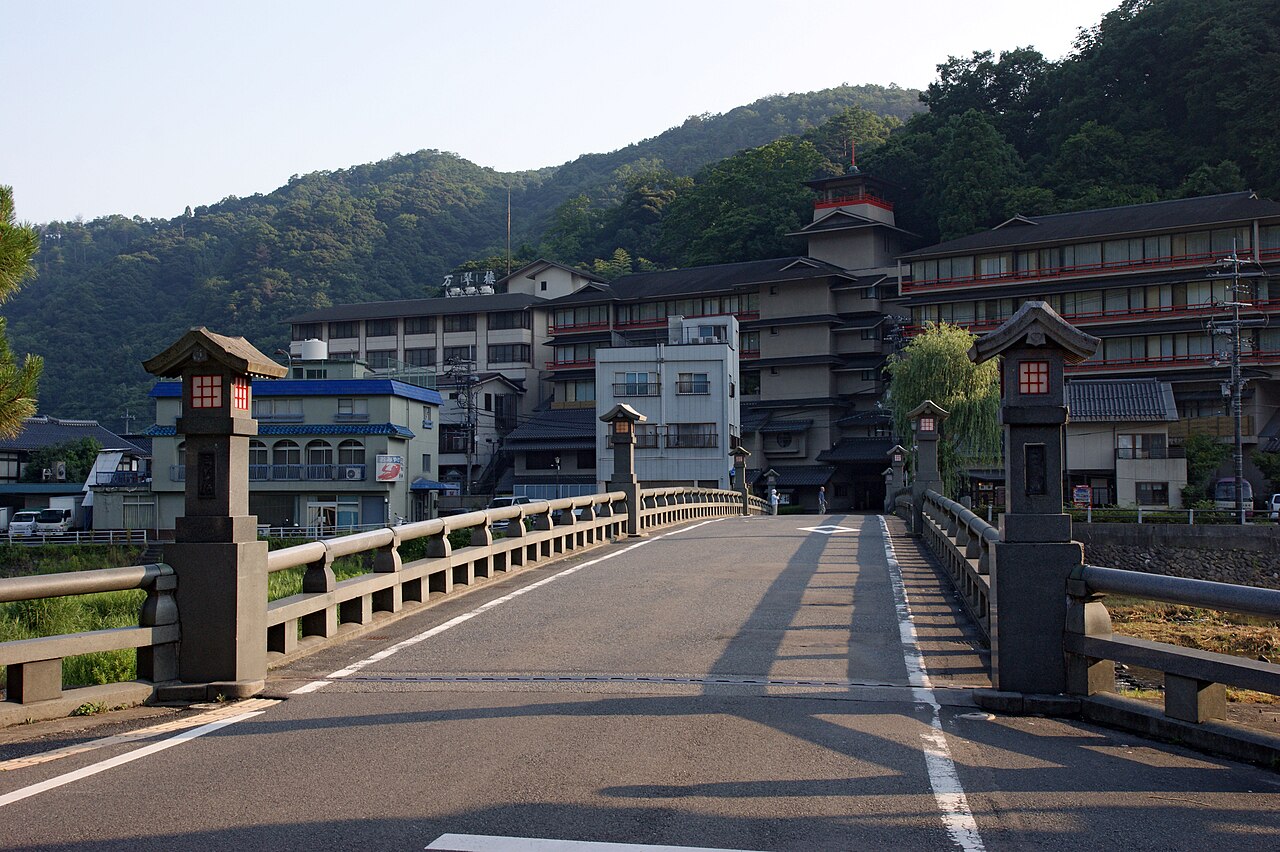 A river bridge with stone lamp posts in Misasa Onsen leading toward a row of ryokan buildings set against forested hills