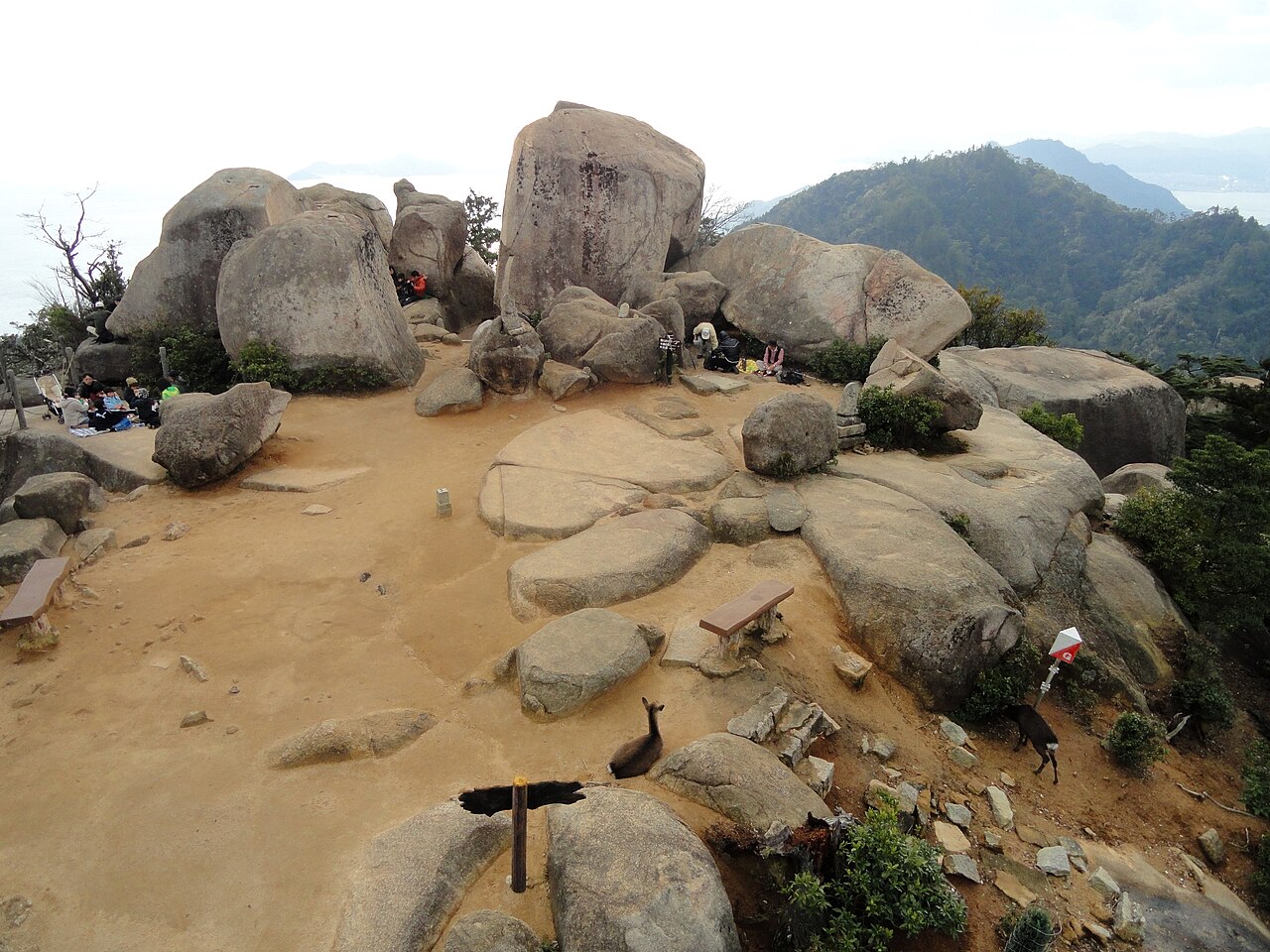 Large granite boulders on the sandy summit of Mount Misen, with forested ridges and a hazy sea beyond and a couple of tiny figures among the rocks.