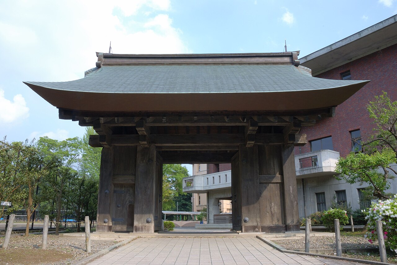 The weathered wooden Yakui Gate of Mito Castle with a broad, curved green copper roof, standing among trees beside a modern school building.