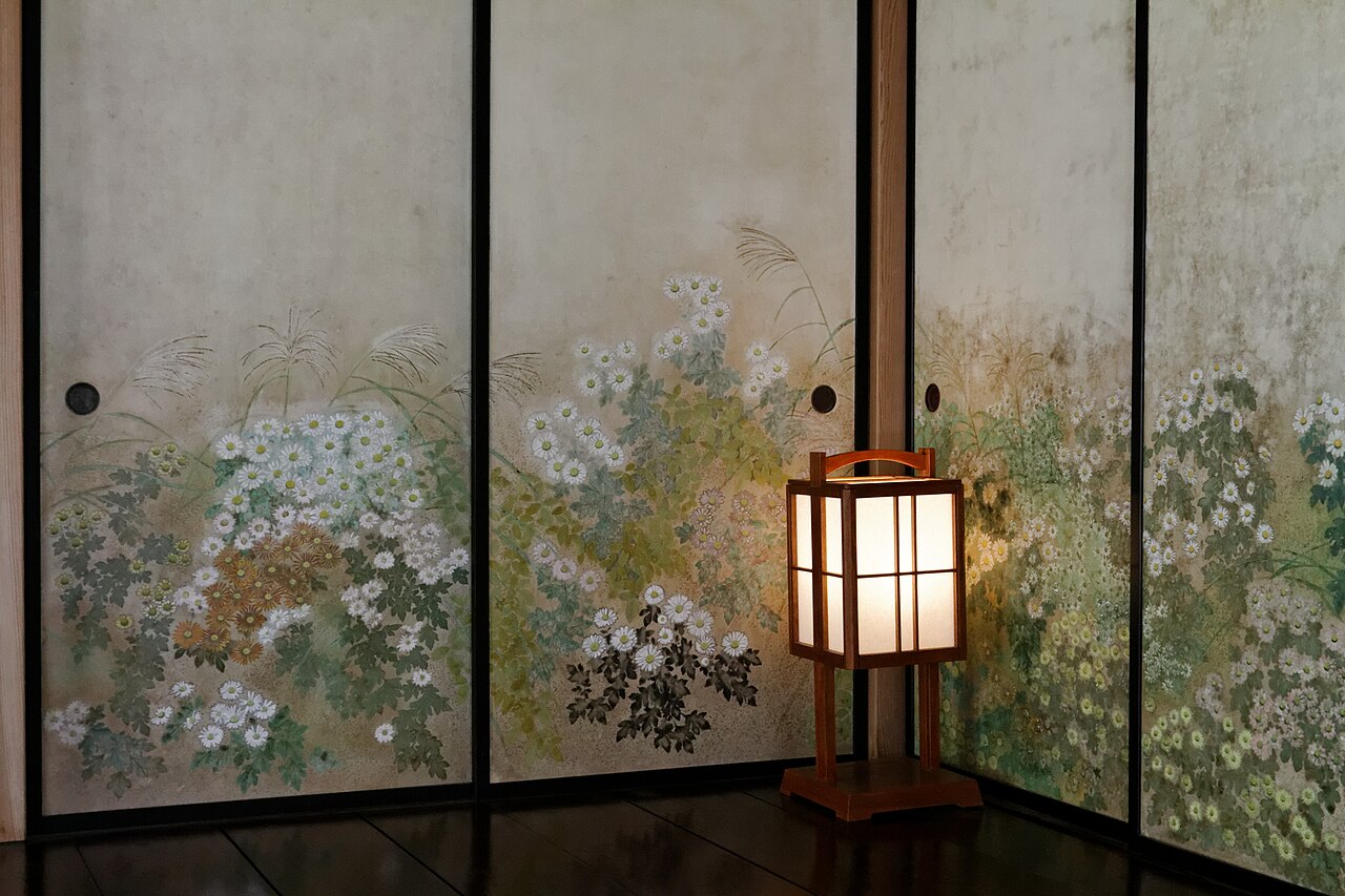 Sliding paper doors painted with white and bronze chrysanthemums and pampas grass, with a lit paper lantern standing in the corner of a room in Kobuntei, Kairakuen.