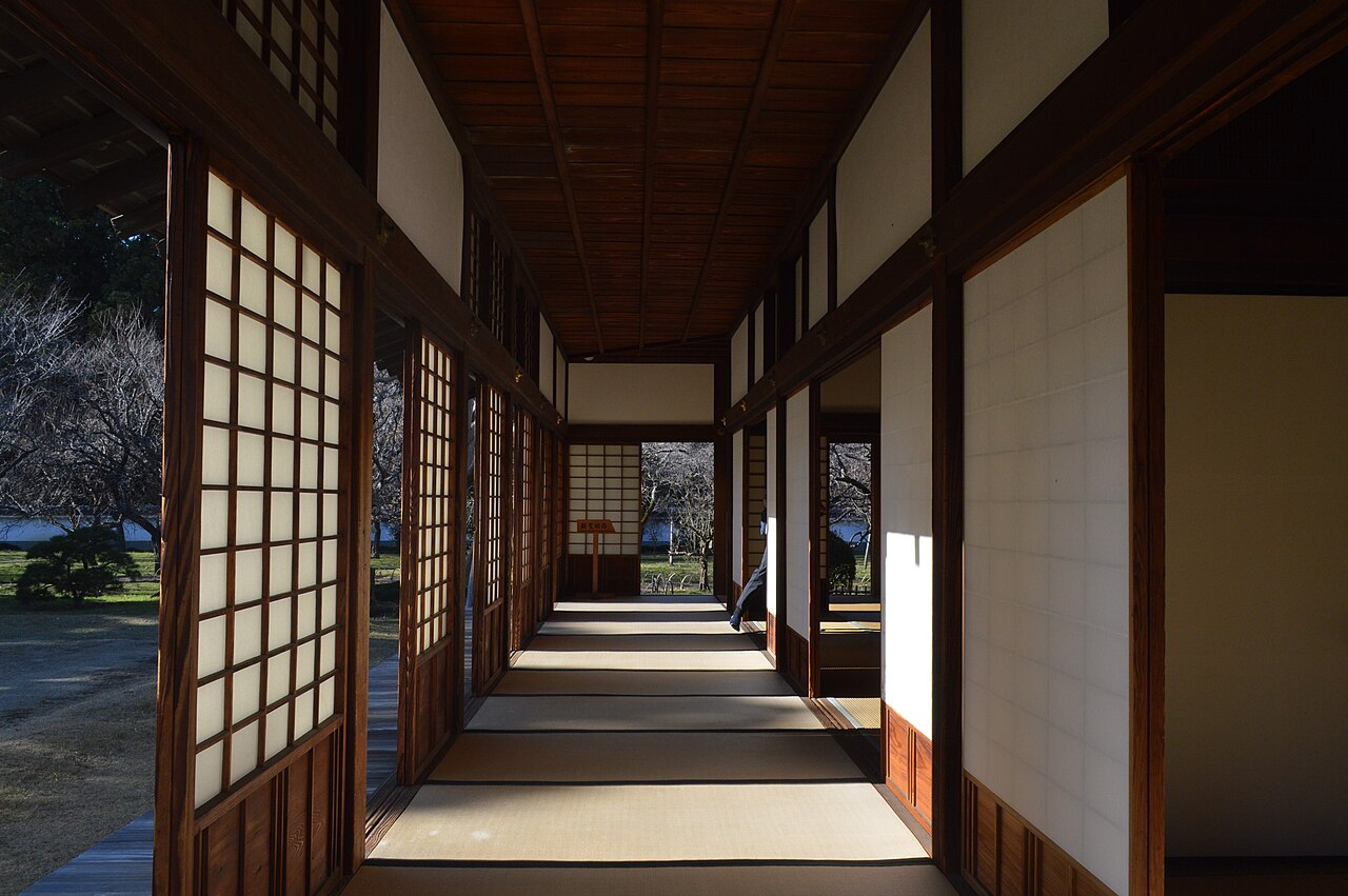 A long tatami corridor lined with paper screens and wooden lattice doors, lit by low sunlight, inside the Kodokan former domain school in Mito.