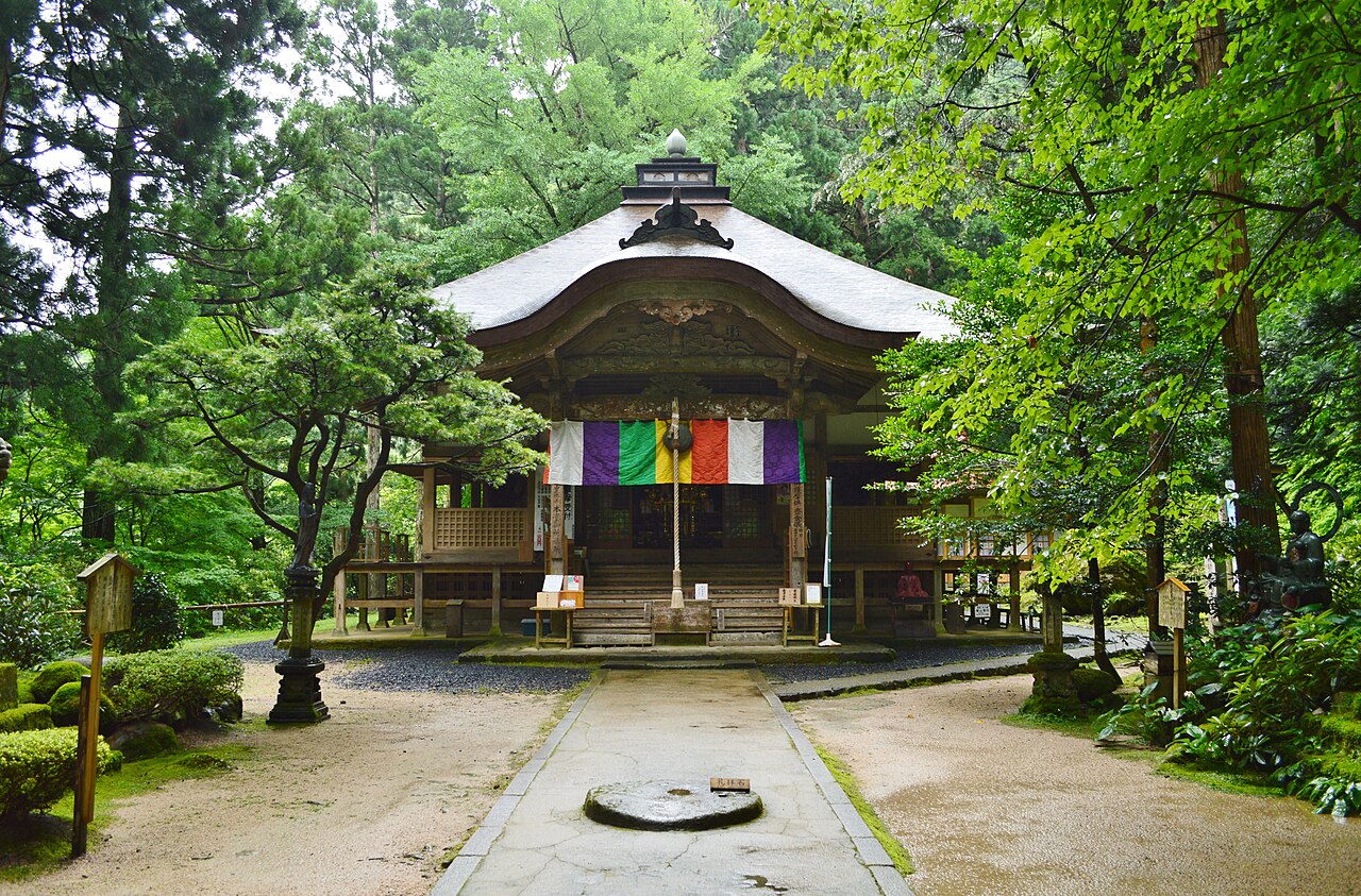 The main hall of Sanbutsu-ji with colorful hanging cloths at its entrance, seen across a gravel courtyard framed by green trees