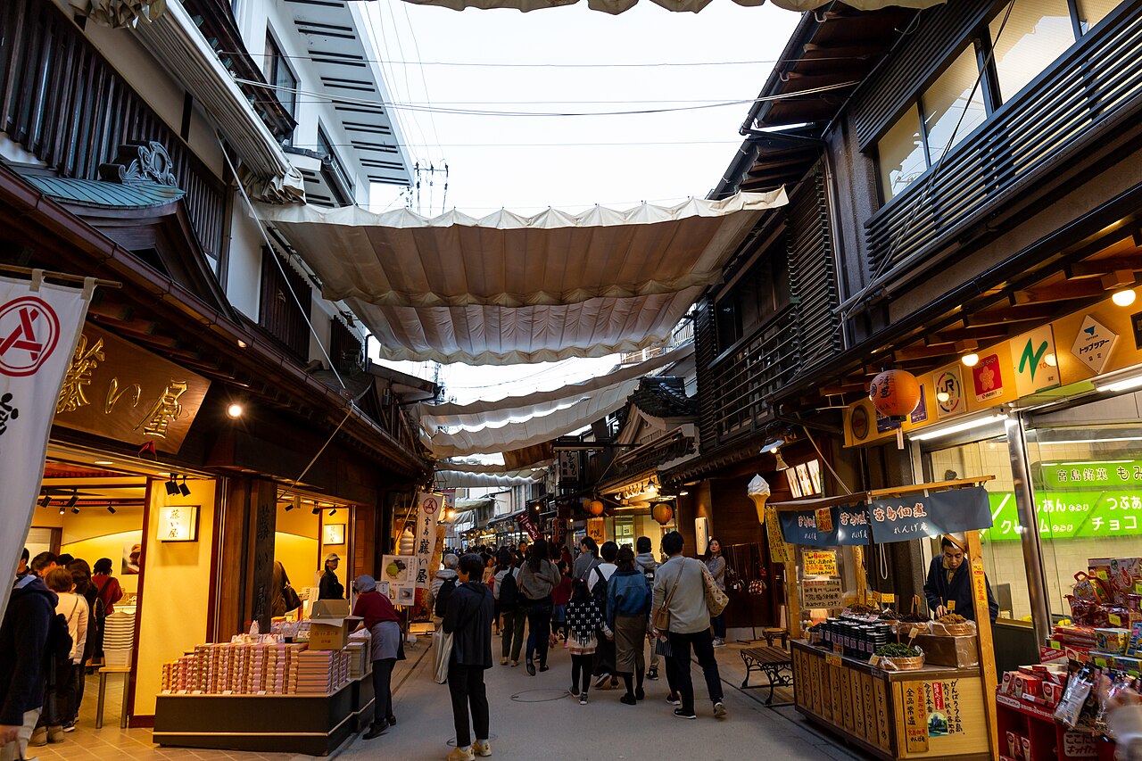 A shopping street on Miyajima lined with wooden shopfronts and snack stalls under pale canvas awnings, with a few visitors walking between them.