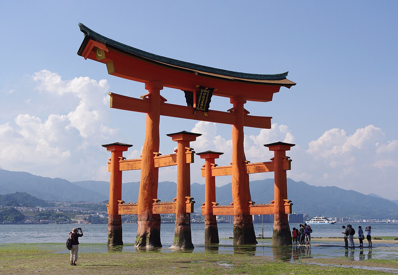 The vermilion great torii of Itsukushima Shrine standing on damp sand at low tide, with hazy mountains and a strip of sea behind it and a few small visitors near its feet.