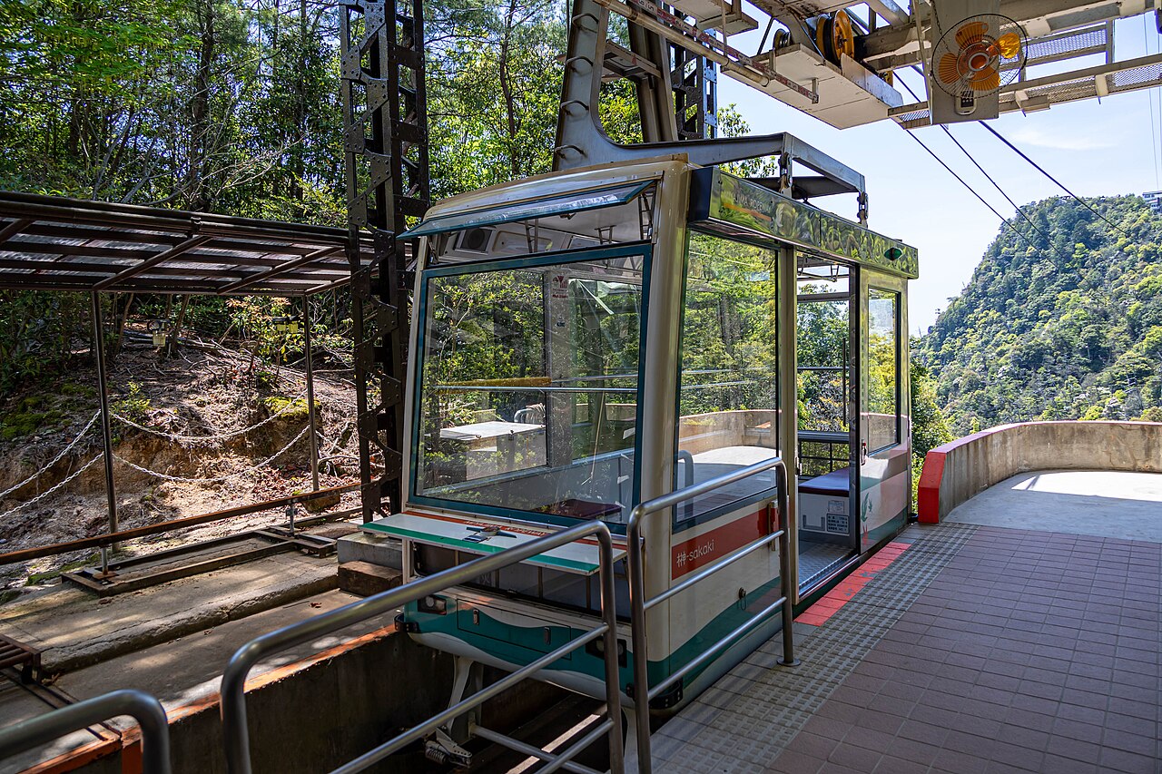 A glass-sided ropeway gondola waiting at an open station platform among steel supports, with steep wooded slopes and a rocky cliff behind.