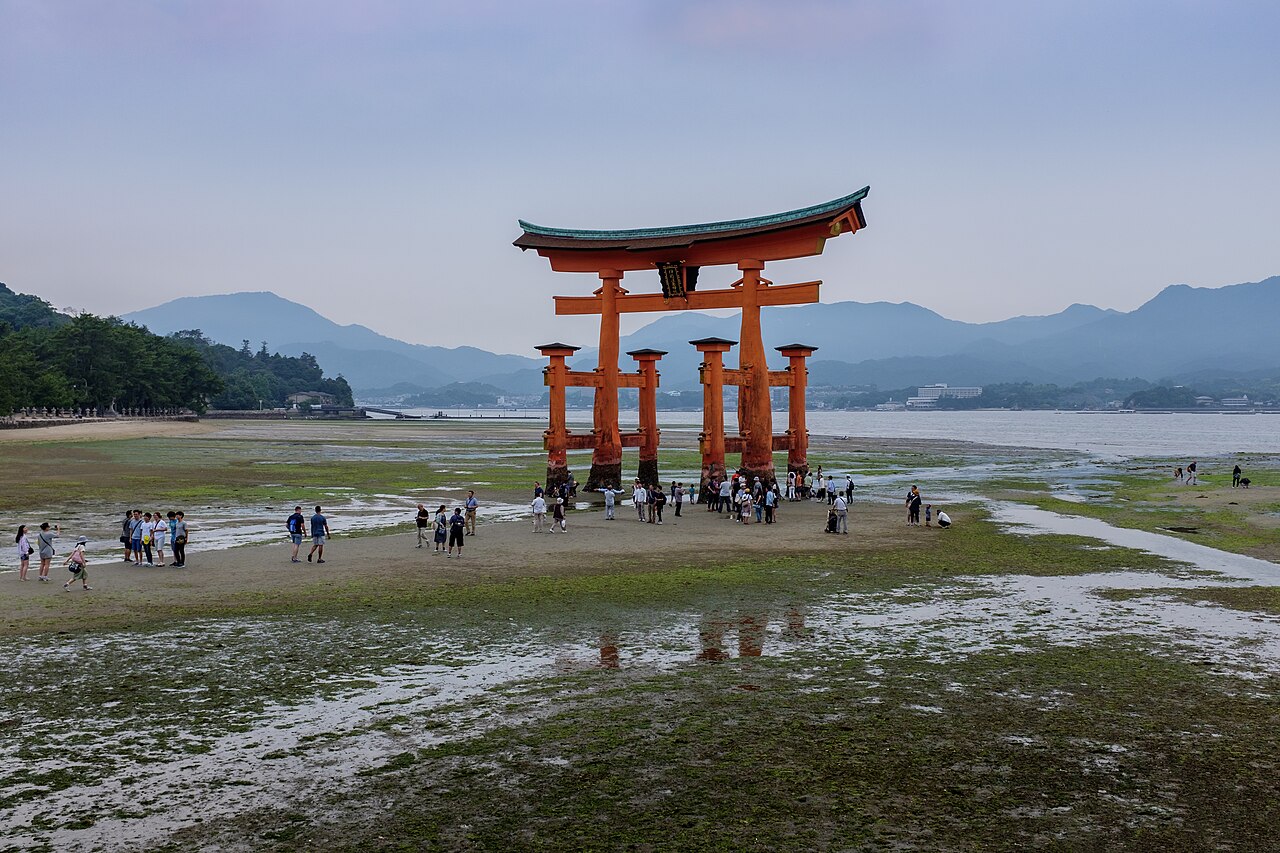 A wide stretch of wet sand and shallow tidal pools at low tide, with the vermilion torii of Itsukushima Shrine in the distance, small figures walking the flats and green mountains behind.