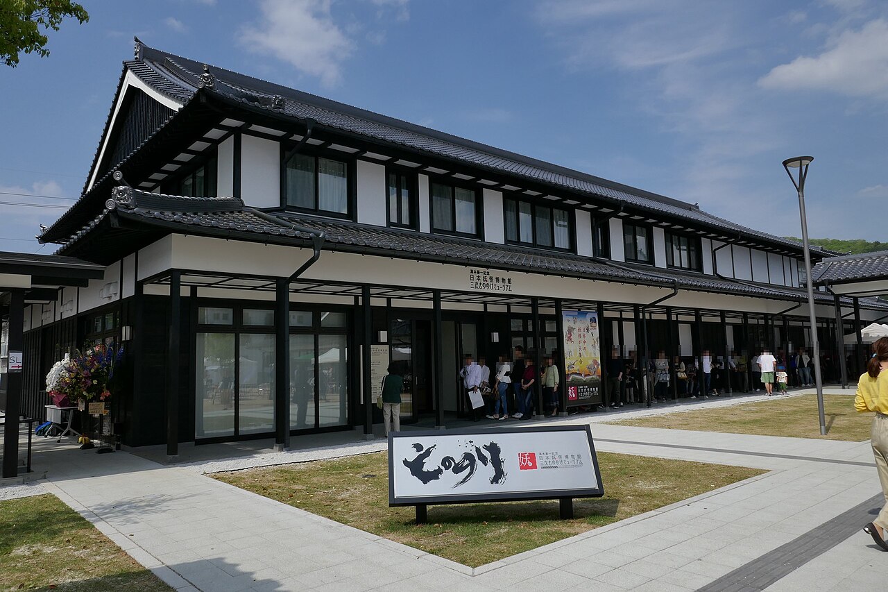A low black-and-white museum building with deep tiled eaves and a large wooden sign in front, seen across a paved forecourt under a blue sky.