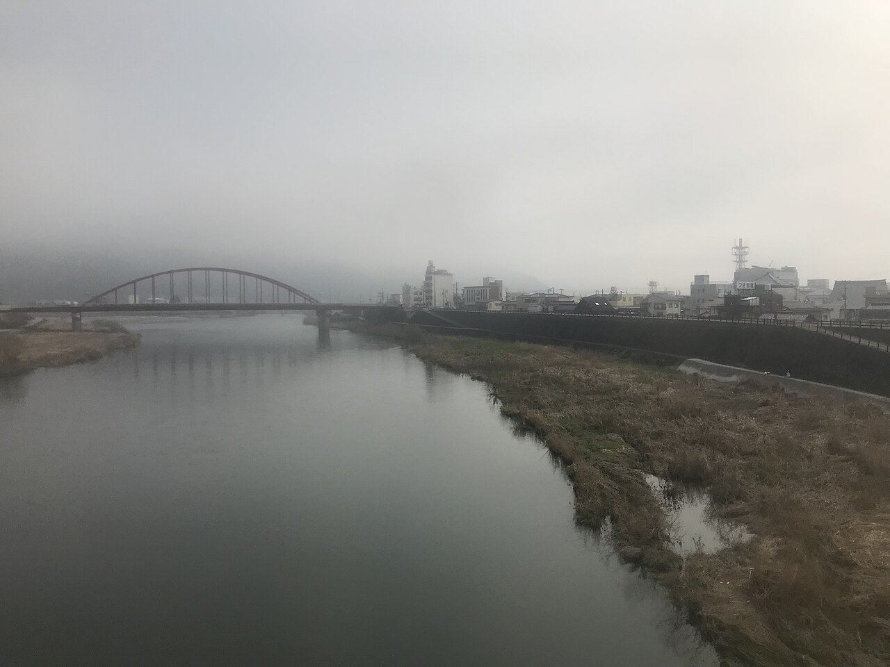 A wide river under a low grey sky with an arched bridge in the mist and the buildings of a small city on the far bank.