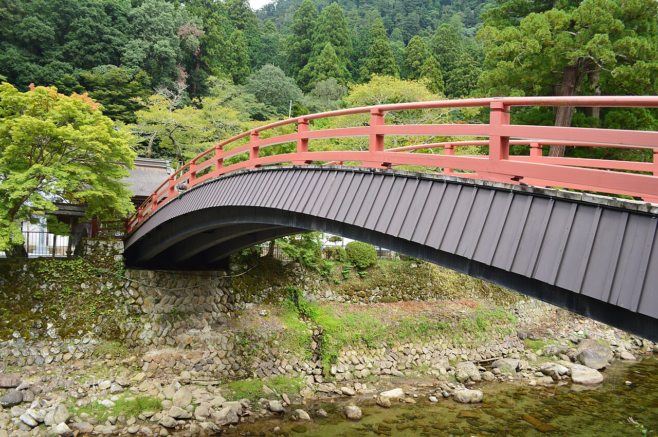A red-railed arched bridge crossing a shallow stony stream below forested slopes.