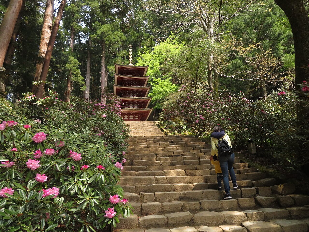 A five-storied red pagoda seen at the top of a wide stone staircase lined with pink rhododendron and tall cedars.