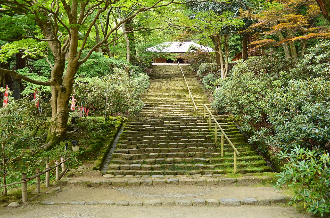 A long moss-covered stone staircase climbing between trees and rhododendron bushes toward a hall at the top.