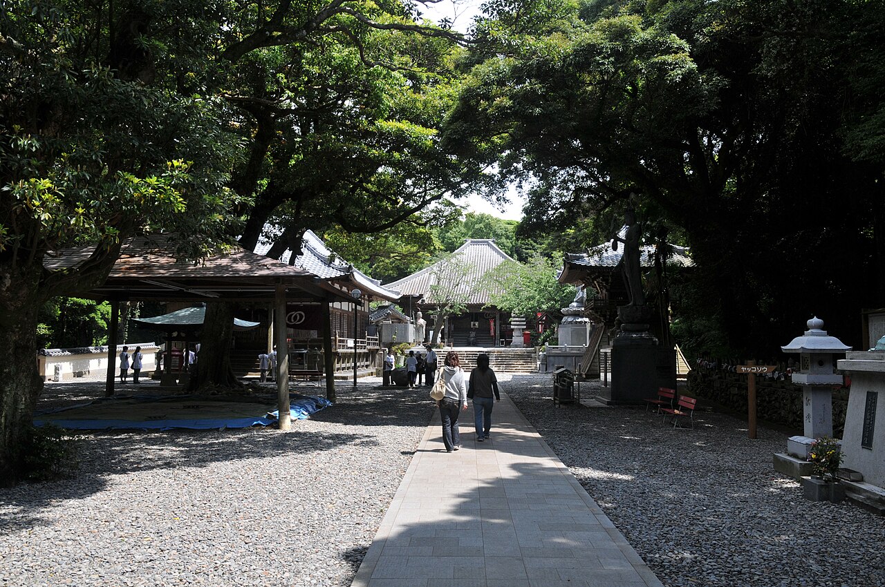 A paved path running between gravel courtyards and tall evergreen trees at Hotsumisaki-ji, with visitors walking toward the temple halls and stone lanterns at the right