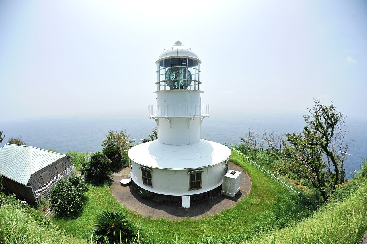 The white Muroto-misaki Lighthouse standing on a grassy cliff top above a hazy Pacific horizon, photographed with a wide lens so the tower and keeper's house curve slightly
